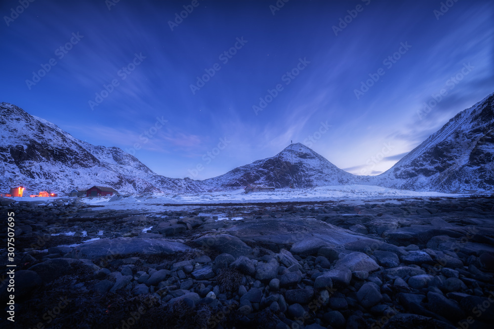 Blue sky with stars and clouds over rocky beach and snow covered mountains at night Lofoten islands,