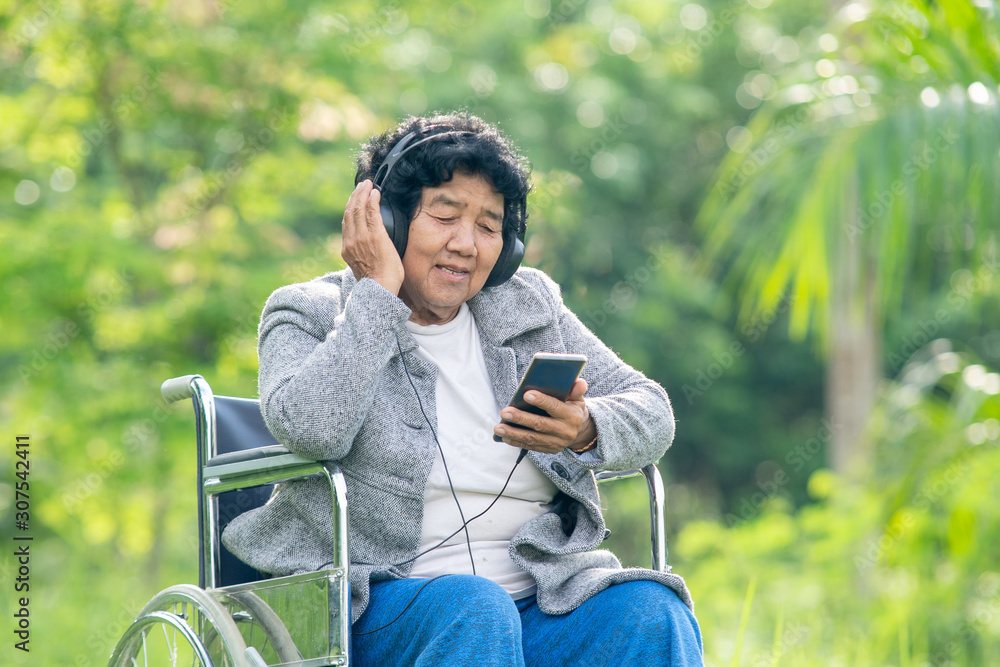 坐在轮椅上的亚洲老年女性在公园里用智能手机听音乐，音乐疗法