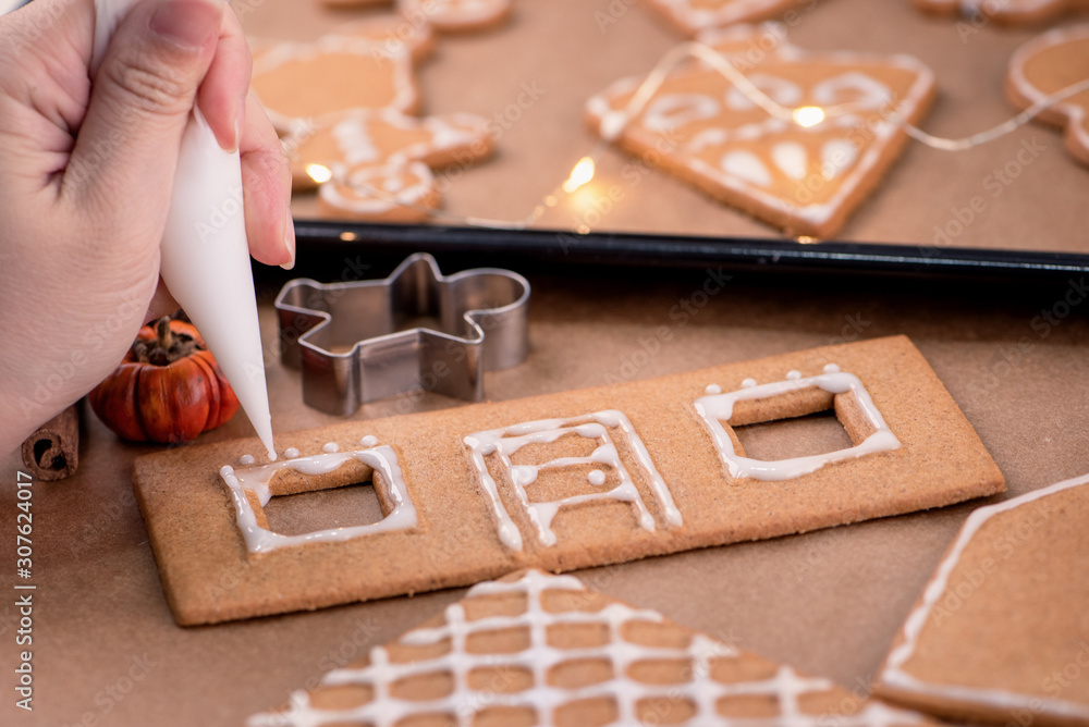 Woman is decorating gingerbread cookies house with white frosting icing cream topping on wooden tabl