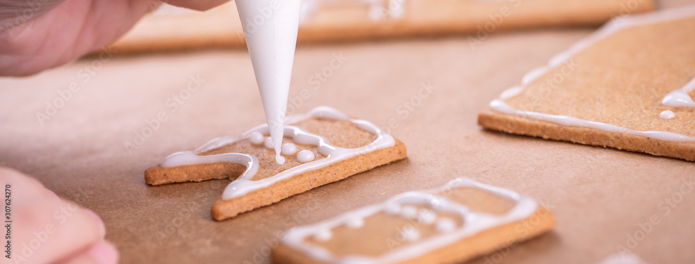 Woman is decorating gingerbread cookies house with white frosting icing cream topping on wooden tabl