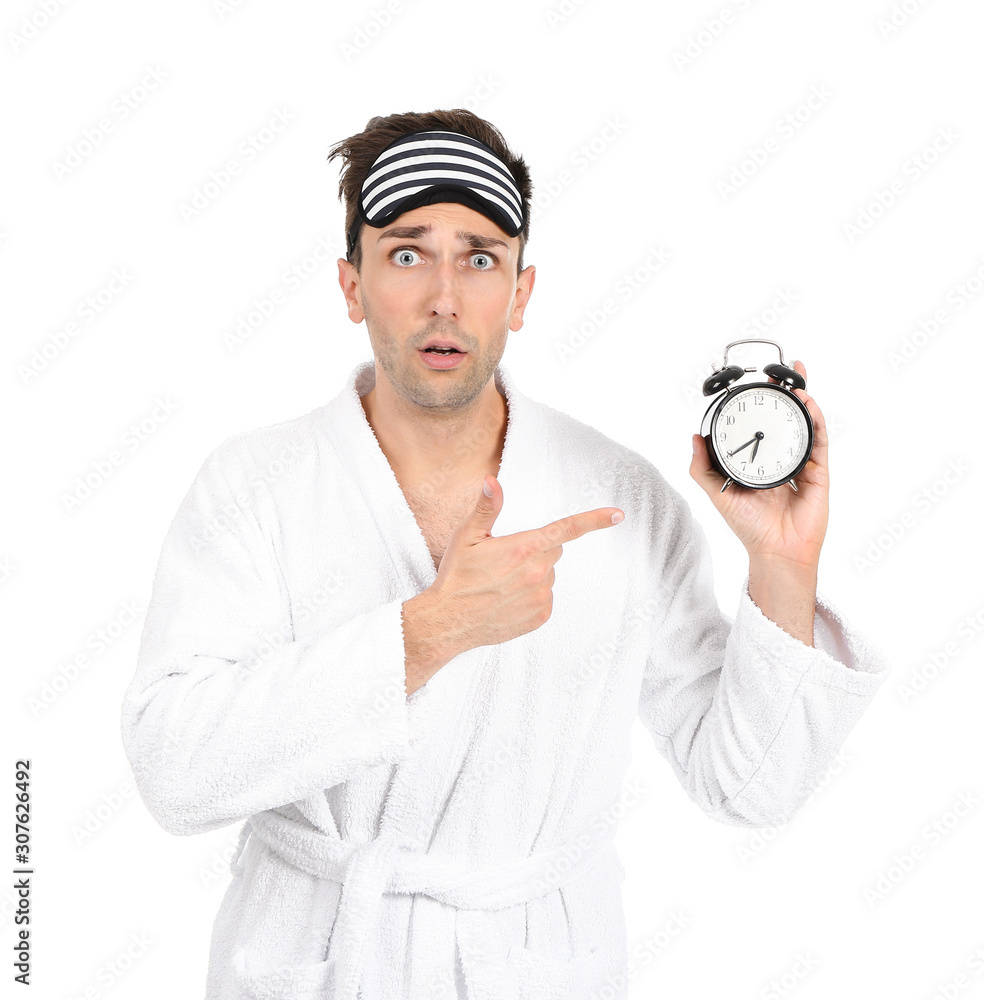 Surprised man in bathrobe, sleep mask and alarm clock on white background