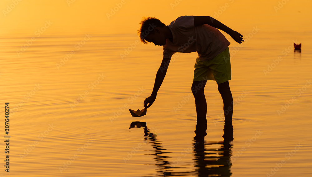 Teen boy launching paper ships on water. Floating ships into the distance. Beautiful orange sunset. 