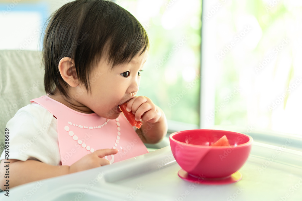 坐在婴儿高脚椅上喂奶的小可爱，在家里面带微笑地抱着西瓜。孩子