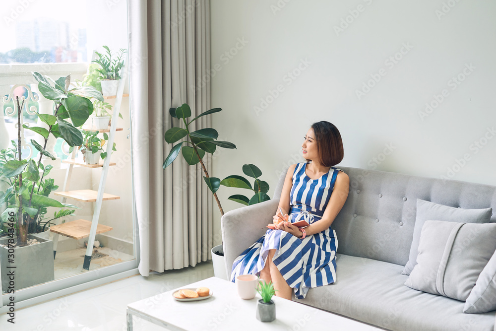 Young beautiful Asian woman writing and sitting on sofa in living room.