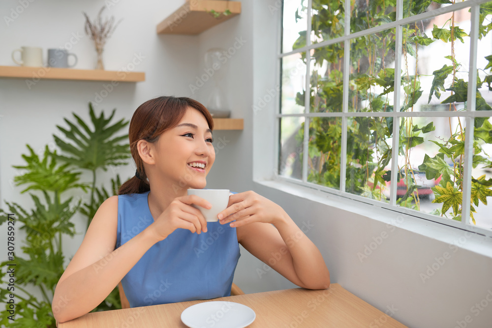 快乐的年轻亚洲女人在家里靠窗喝咖啡