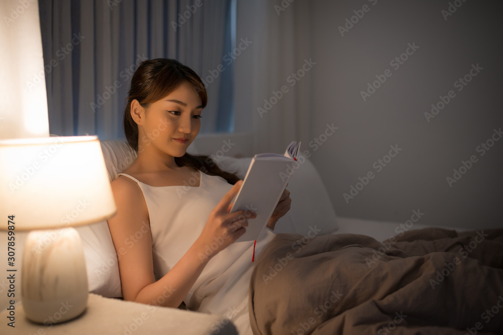 leisure and people concept - young woman reading book in bed at night home