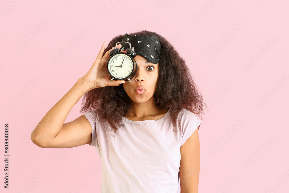 Surprised African-American woman with sleep mask and alarm clock on color background