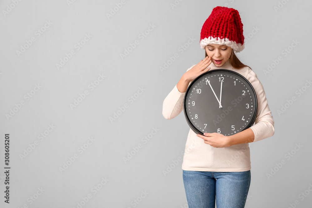 Surprised young woman in Santa hat and with clock on light background. Christmas countdown concept