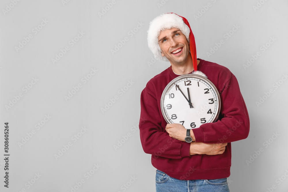 Young man in Santa hat and with clock on light background. Christmas countdown concept