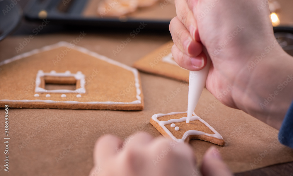 Woman is decorating gingerbread cookies house with white frosting icing cream topping on wooden tabl