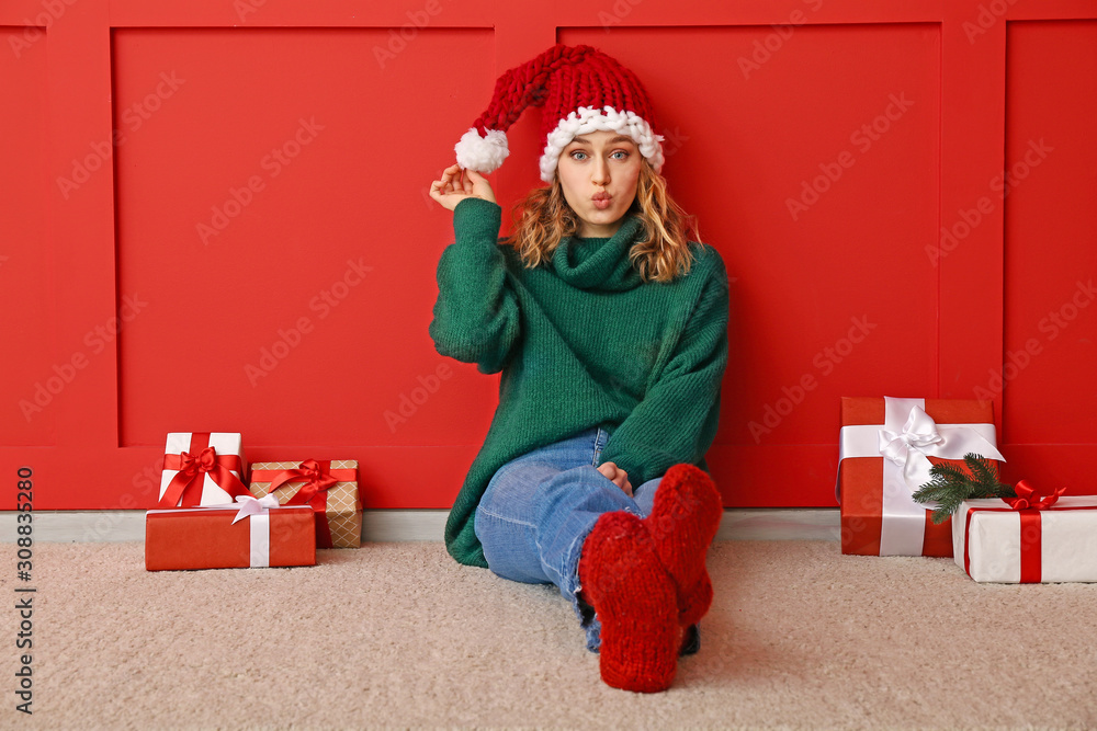 Happy young woman in winter clothes and with Christmas gifts sitting near color wall