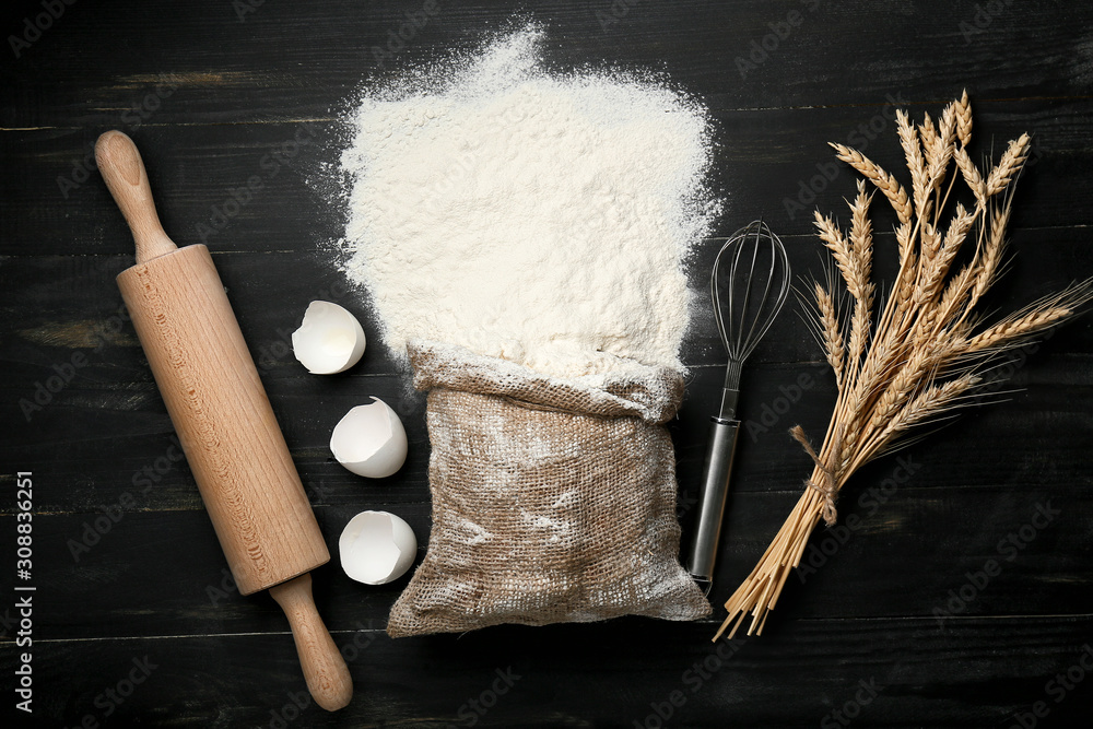 Bag with flour, utensils, egg shell and wheat spikelets on dark background