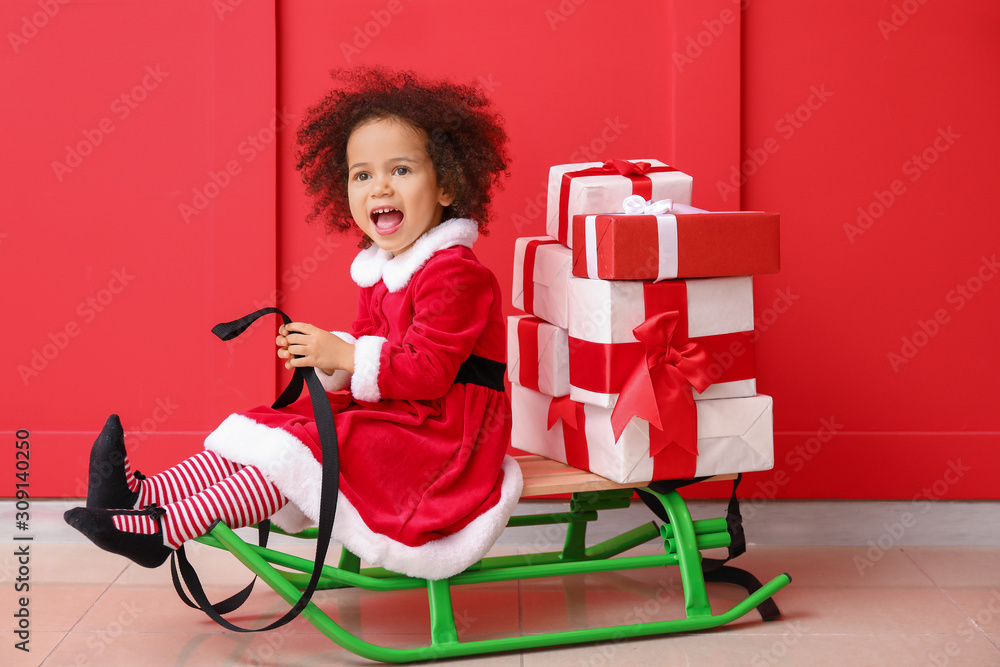 Little African-American girl in Santa costume sitting on sledges with gift boxes near color wall