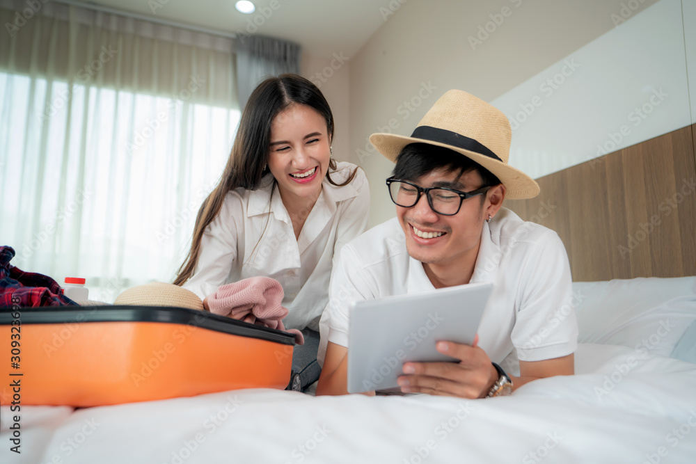 Happy couple packing suitcase on bed in bedroom and lying and looking digital tablet for search and 