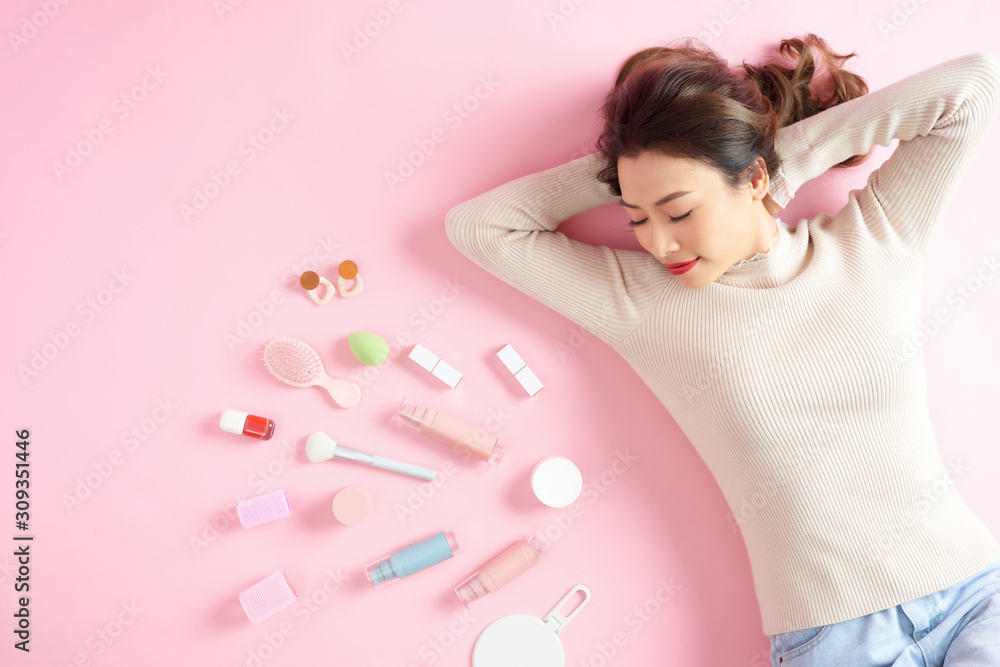 Sleeping Asian woman lying on pink background with her cosmetics. Top view