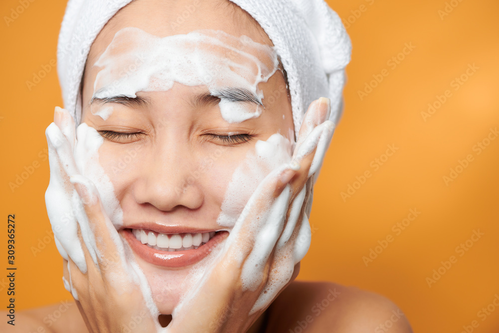 Gentle half-naked woman wrapped in towel washing her face with foaming cleanser isolated over orange