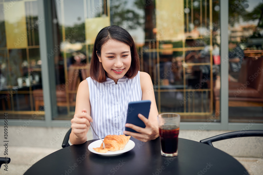 Smiling pretty young woman in casual shirt sitting at table in cafe and checking messenger while usi