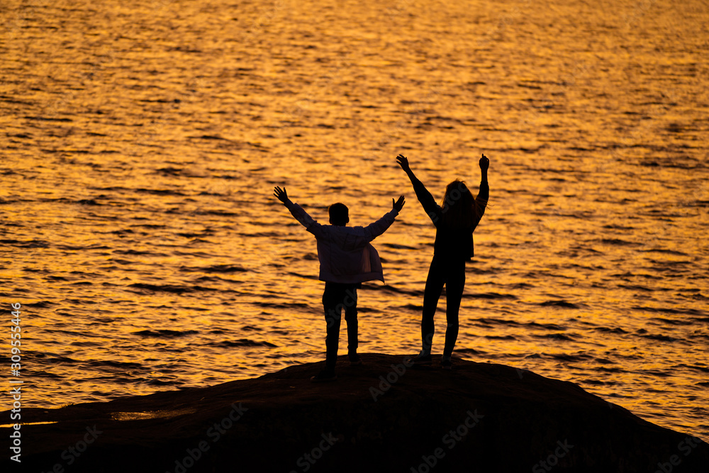 Two young people standing on rock in front of water with hands up looking at sundown. silhuette agai