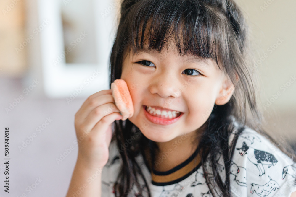 Lovely little asian girl is applying the make up on her cheek with special brush on and looking mirr