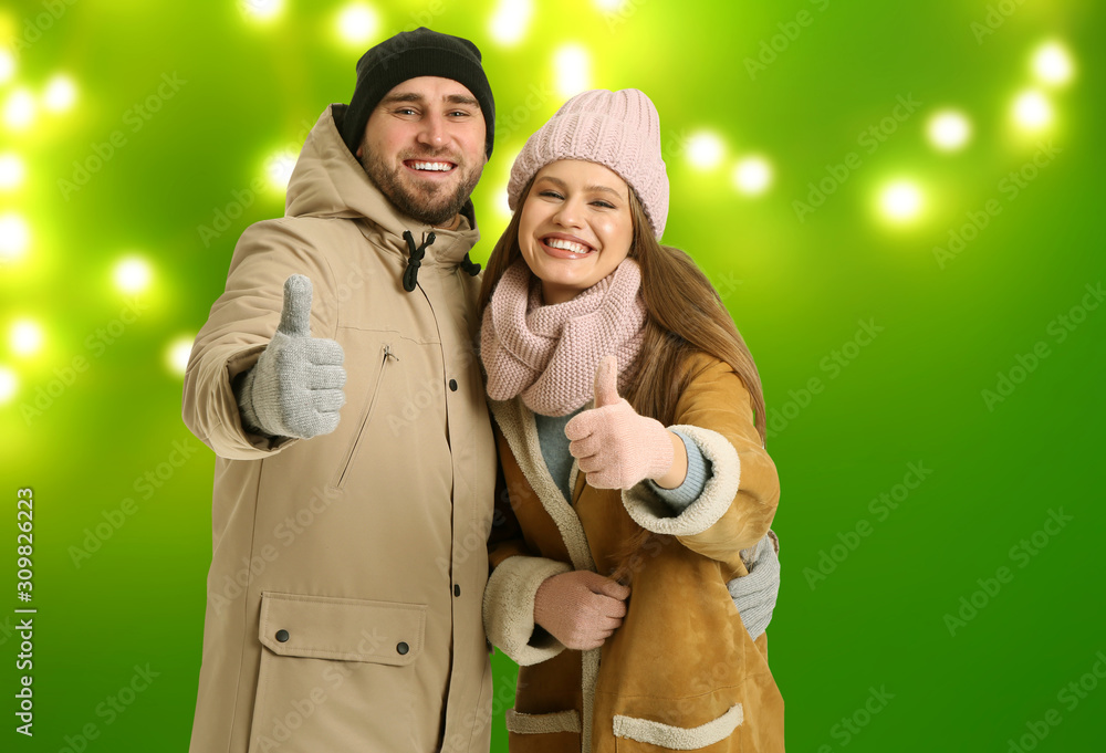 Portrait of happy couple in winter clothes showing thumb-up gesture on color background with blurred