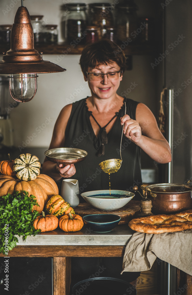 Eldery woman in dark dress serving Autumn seasonal pumpkin cream soup in modern kitchen interior. Fa