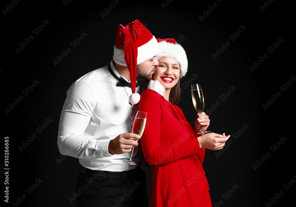 Happy couple in Santa Claus hats and with glasses of champagne on dark background