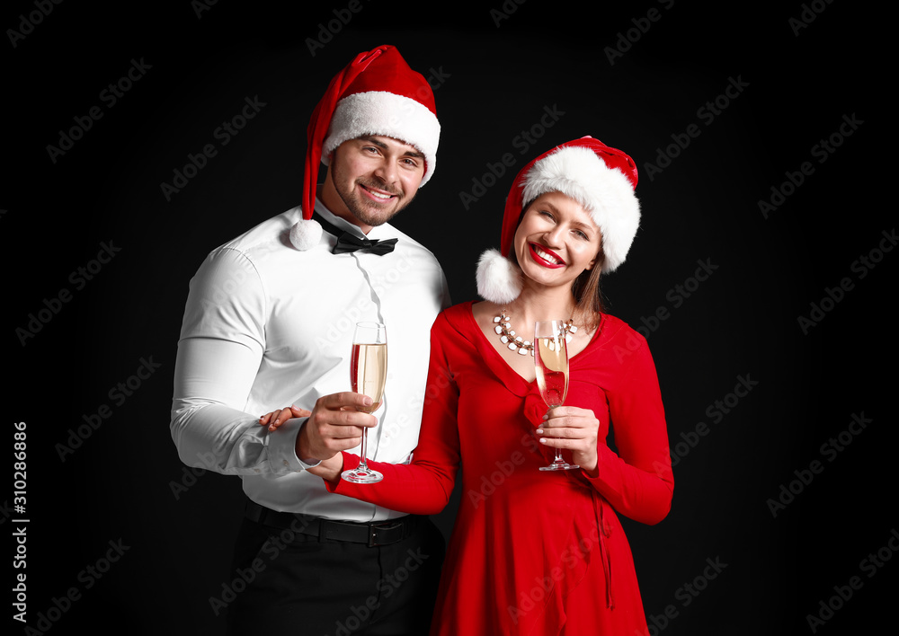 Happy couple in Santa Claus hats and with glasses of champagne on dark background