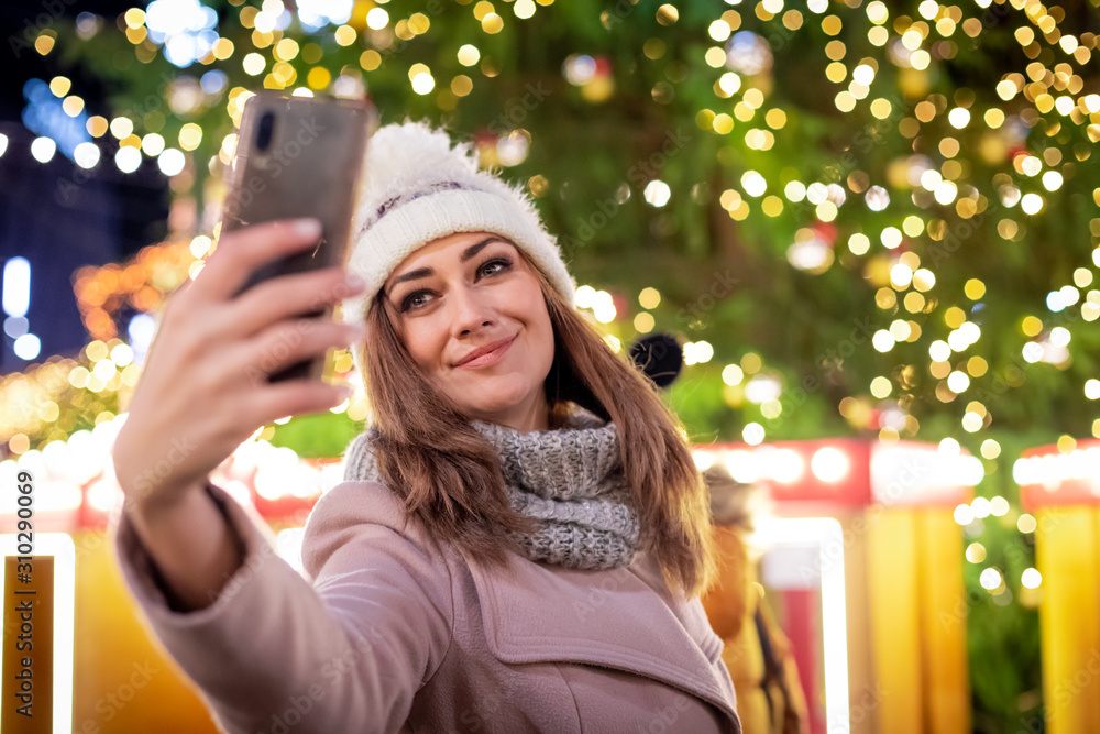 Beautiful woman taking a selfie against the backdrop of a Christmas tree on a decorated street