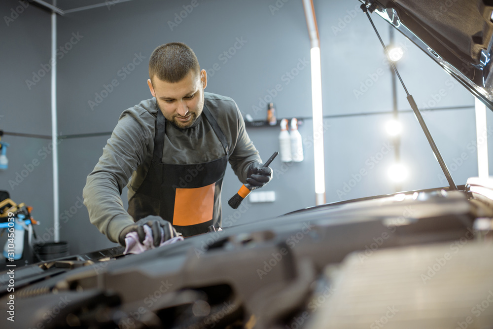 Service worker in uniform performs a professional car cleaning, wiping engine at the vehicle service