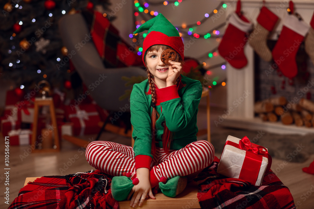 Little girl in costume of elf, with gift and cookie in room decorated for Christmas