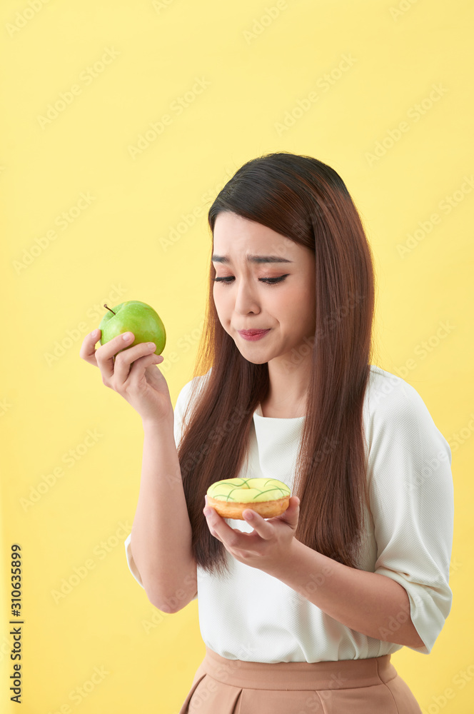 Portrait of a smiling young asian woman choosing between donut and green apple isolated over yellow 