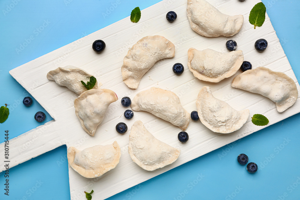 Board with raw dumplings and blueberries on color background