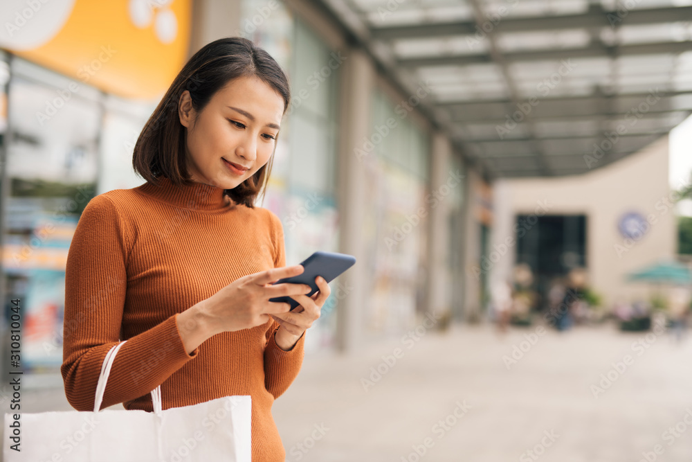 Portrait of elegant young Asian woman holding shopping bags and using smartphone on the go while lea