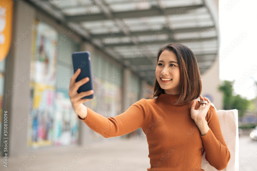 美丽的亚洲年轻女子手持手机与购物袋在户外购物中心自拍