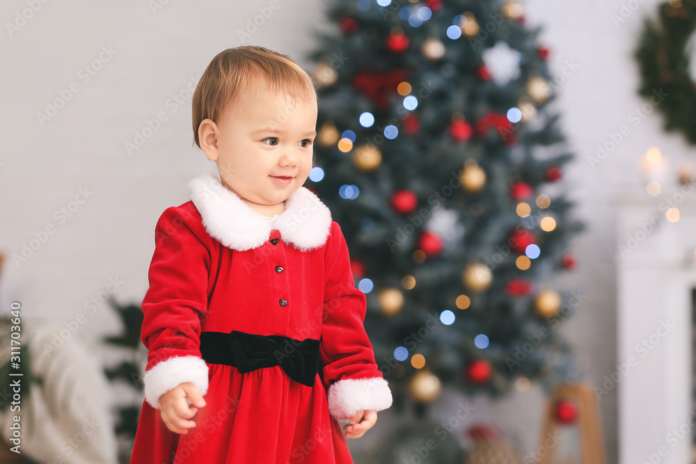 Cute little baby in Santa Claus costume in room decorated for Christmas