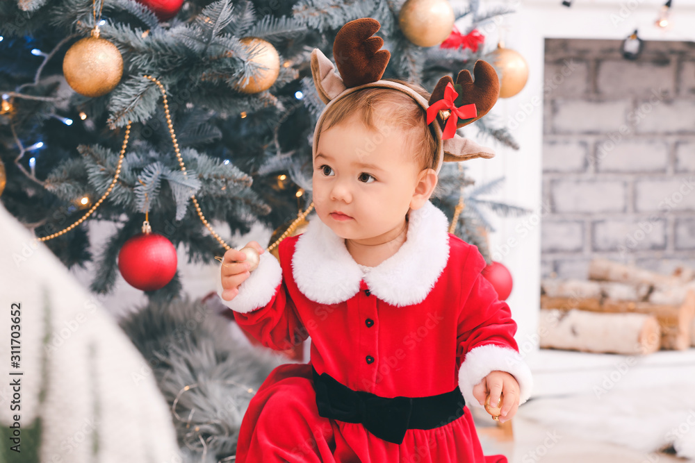 Cute little baby in Santa Claus costume near Christmas tree at home