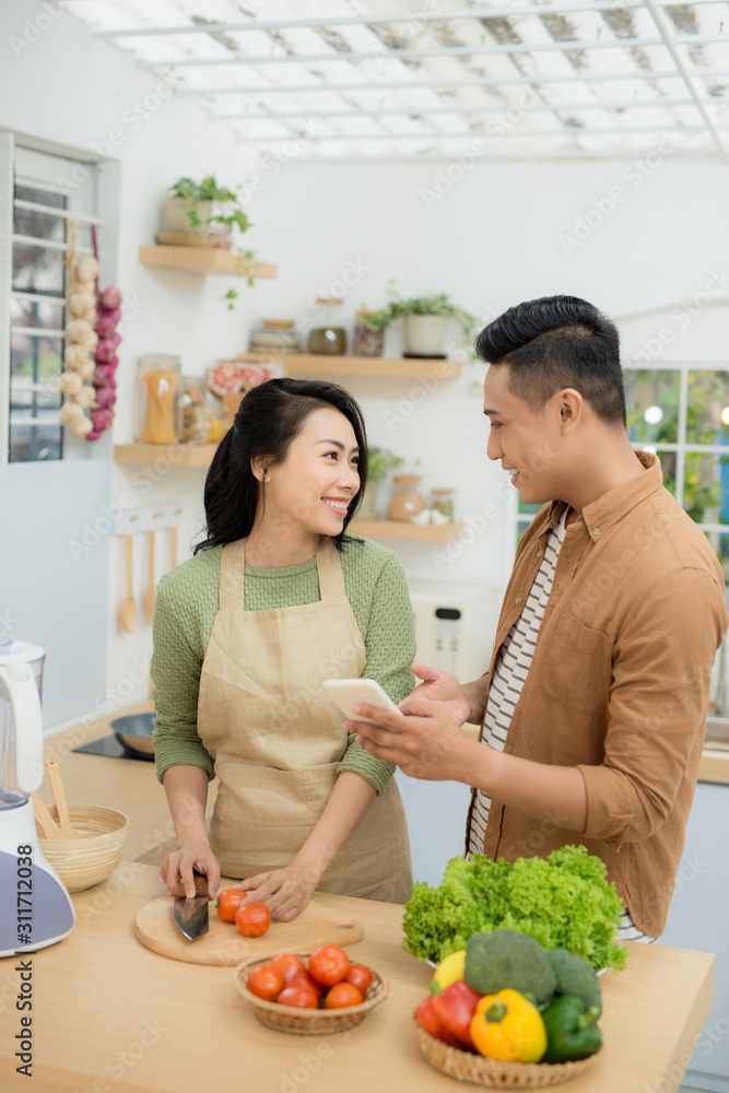 漂亮的年轻夫妇在家里的厨房里做饭时，正在用手机微笑