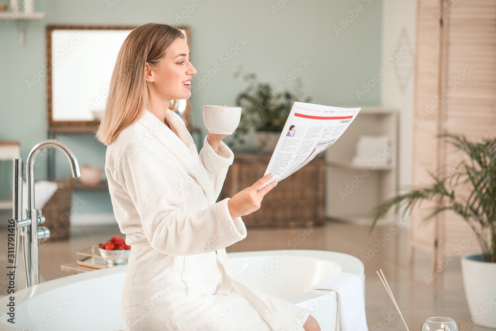 Beautiful young woman drinking tea and reading newspaper in bathroom