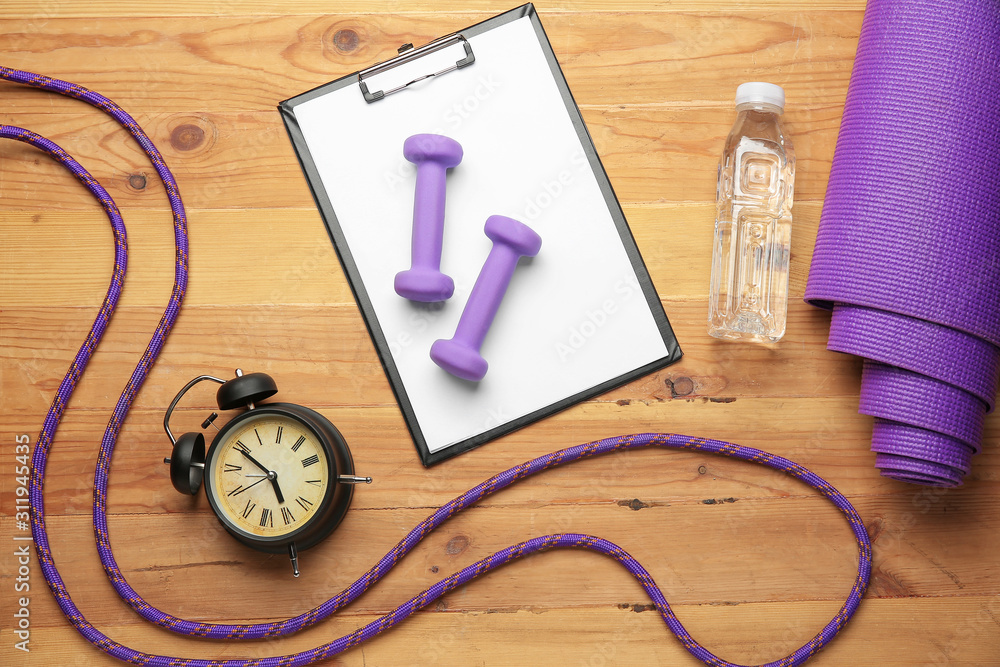 Dumbbells with clipboard, alarm clock, yoga mat, skipping rope and bottle of water on wooden backgro