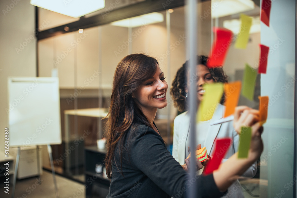 Positive business women working with sticky notes on a glass wall, portrait.