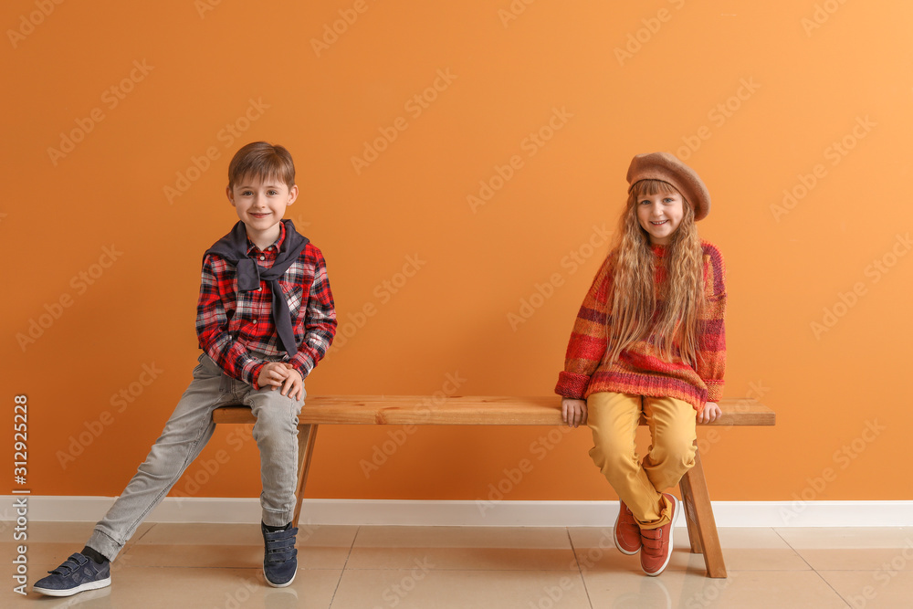 Cute little children in autumn clothes sitting on bench near color wall