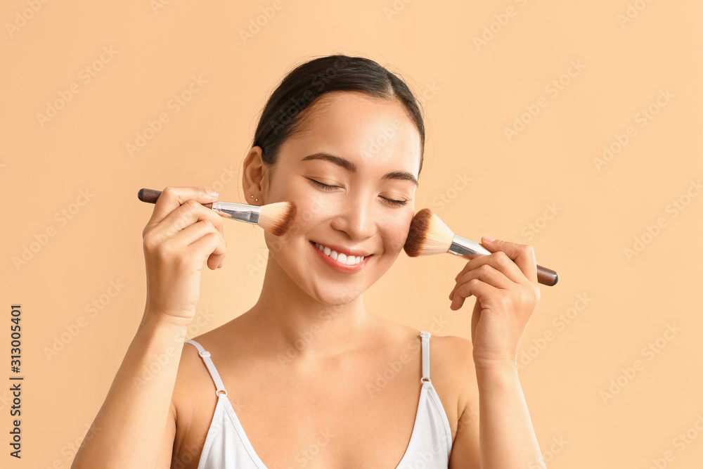 Portrait of young Asian woman with makeup brushes on color background