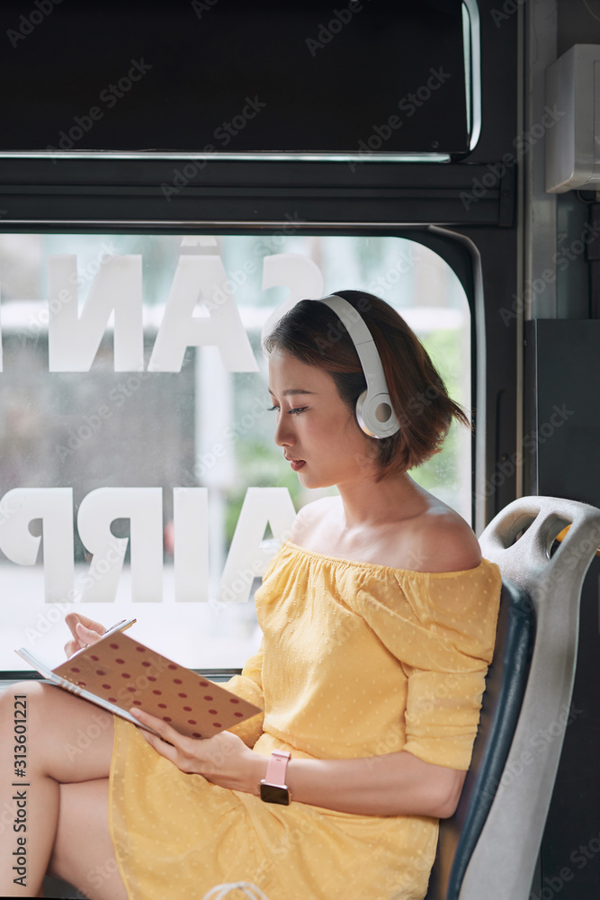 Beautiful young woman sitting in city bus and writing some notes in notebook.