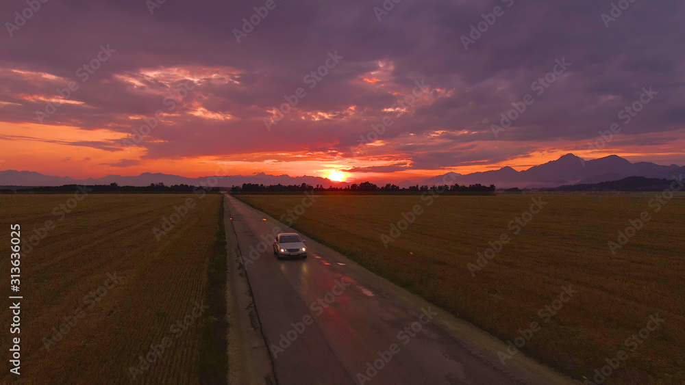 航空：银色汽车在令人惊叹的日落时分沿着风景优美的乡村公路行驶。