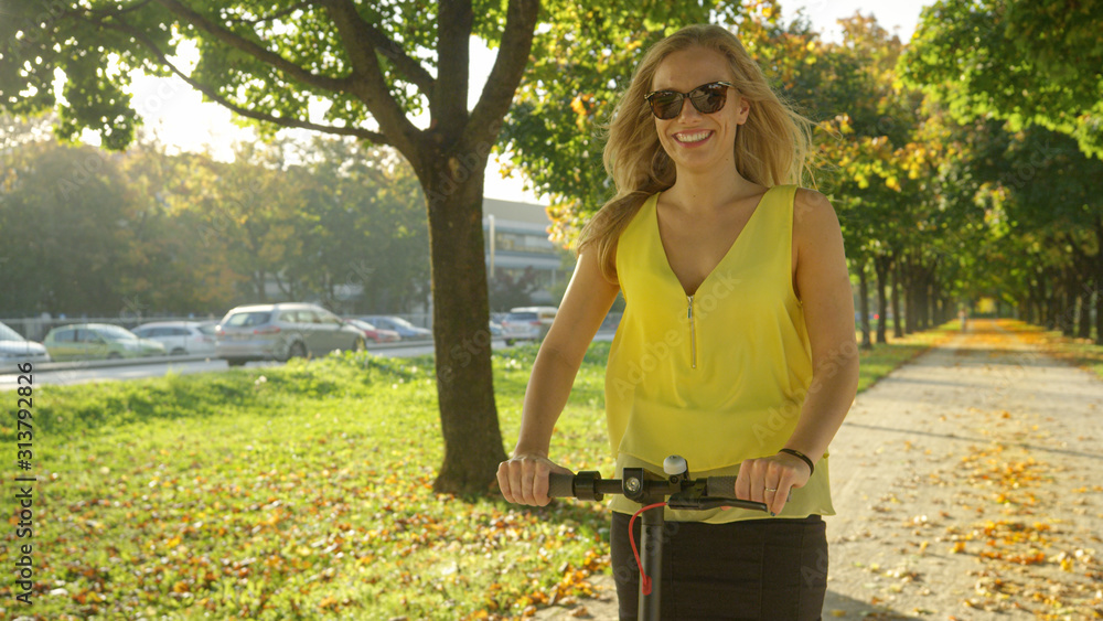 PORTRAIT: Beautiful woman enjoys an electric scooter ride along a sunlit avenue.