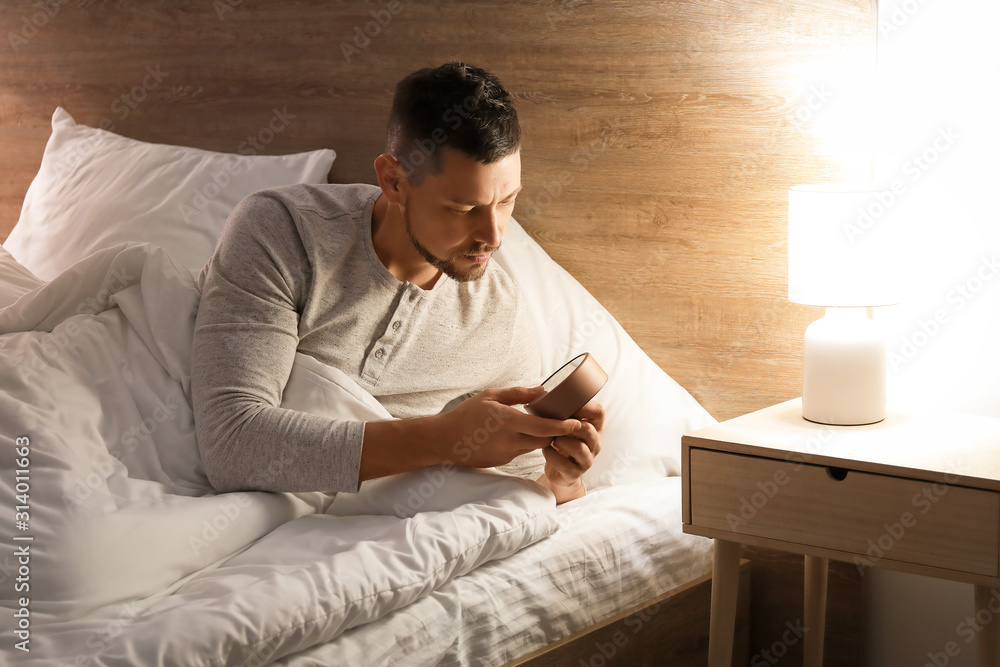 Handsome man setting an alarm while lying in bed at night