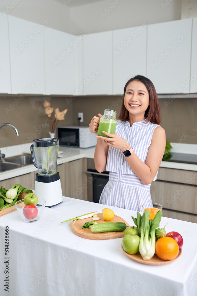 家里厨房里拿着绿色奶昔，面带微笑的年轻亚洲女人。