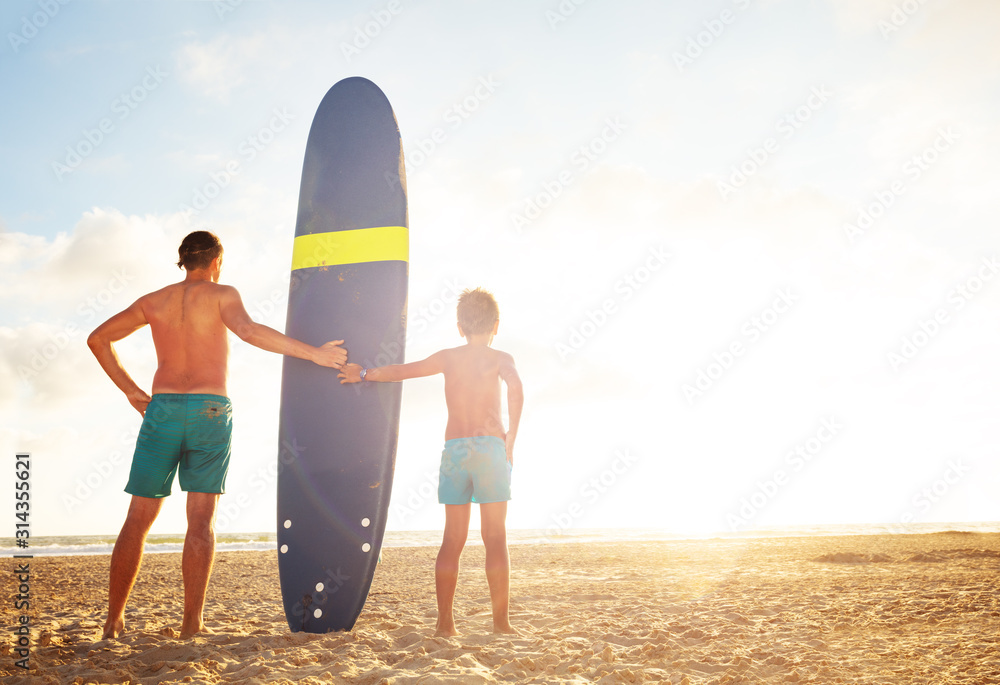 Father and son stand with surf board over sunset