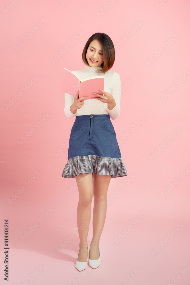 Young asian girl standing, reading interdting book.Isolated on pink background