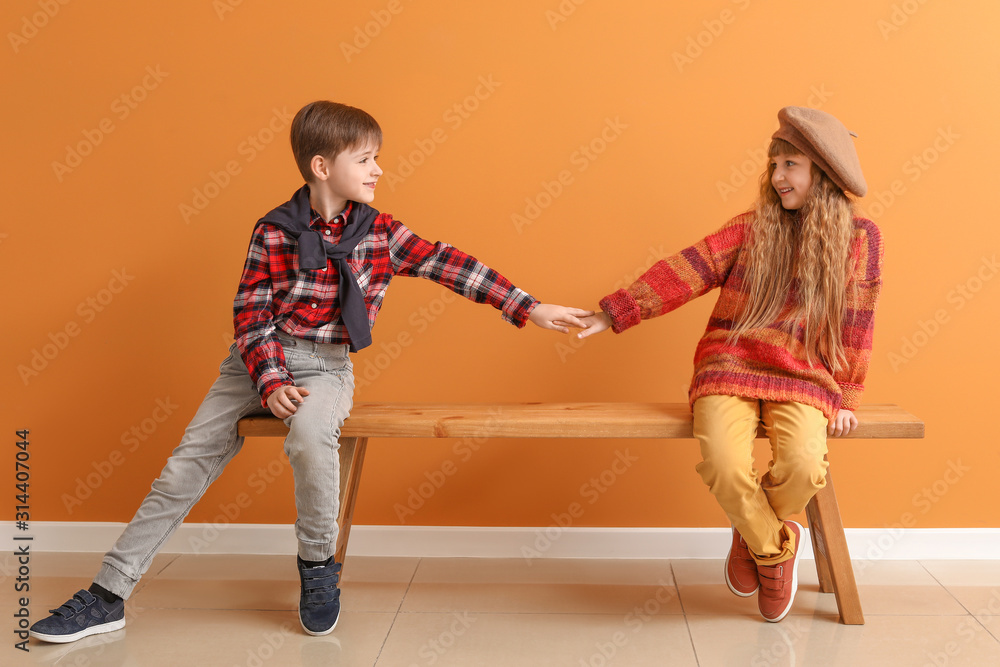 Cute little children in autumn clothes sitting on bench near color wall